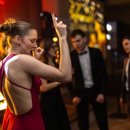Tomasz Gościewicz Fotografia - Kobieta w czerwonej sukni tańczy na parkiecie podczas eventu. W tle inni goście w strojach wieczorowych i dekoracje świetlne.