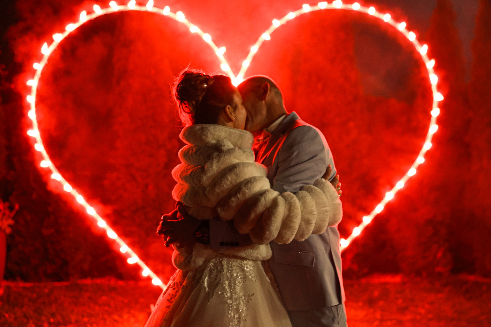 Para młoda w objęciach na tle czerwonego serca z lampek. Panna młoda w białej sukni i futrzanym bolerku, pan młody w jasnym garniturze. Romantyczna atmosfera.