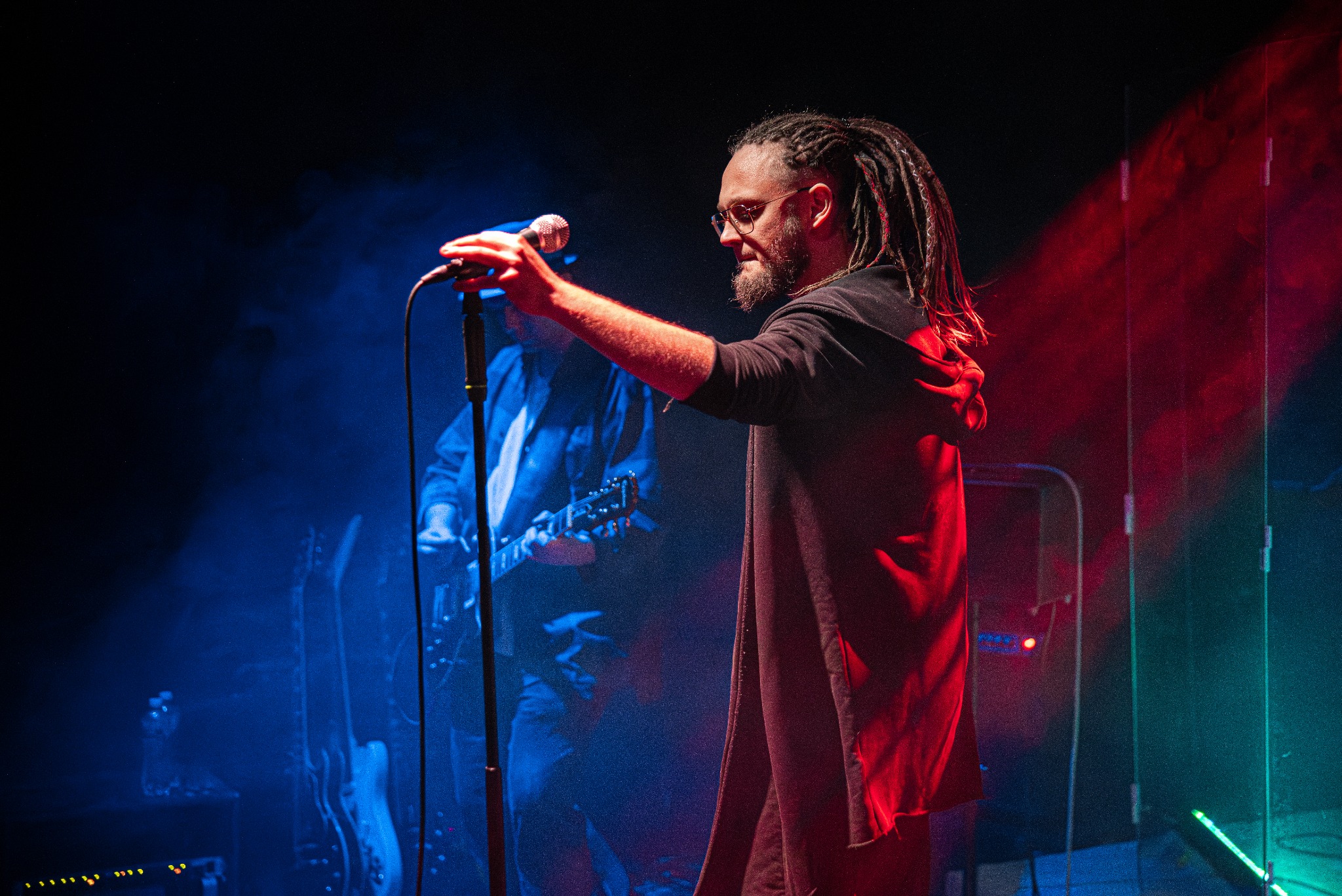 Wokalista z dredami śpiewa do mikrofonu na scenie w czerwono-niebieskim oświetleniu, w tle gitarzysta. Ujęcie z koncertu, dynamiczne światło.