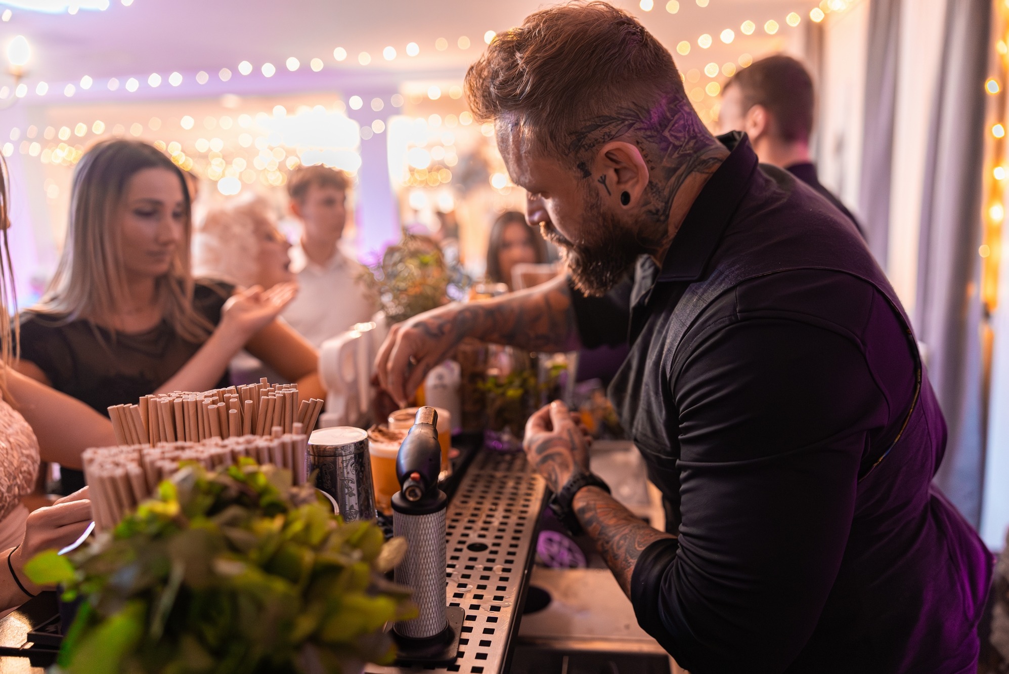Barman z tatuażami przygotowuje drinki na przyjęciu, w tle goście i dekoracyjne światła. Profesjonalna obsługa barmańska na evencie.