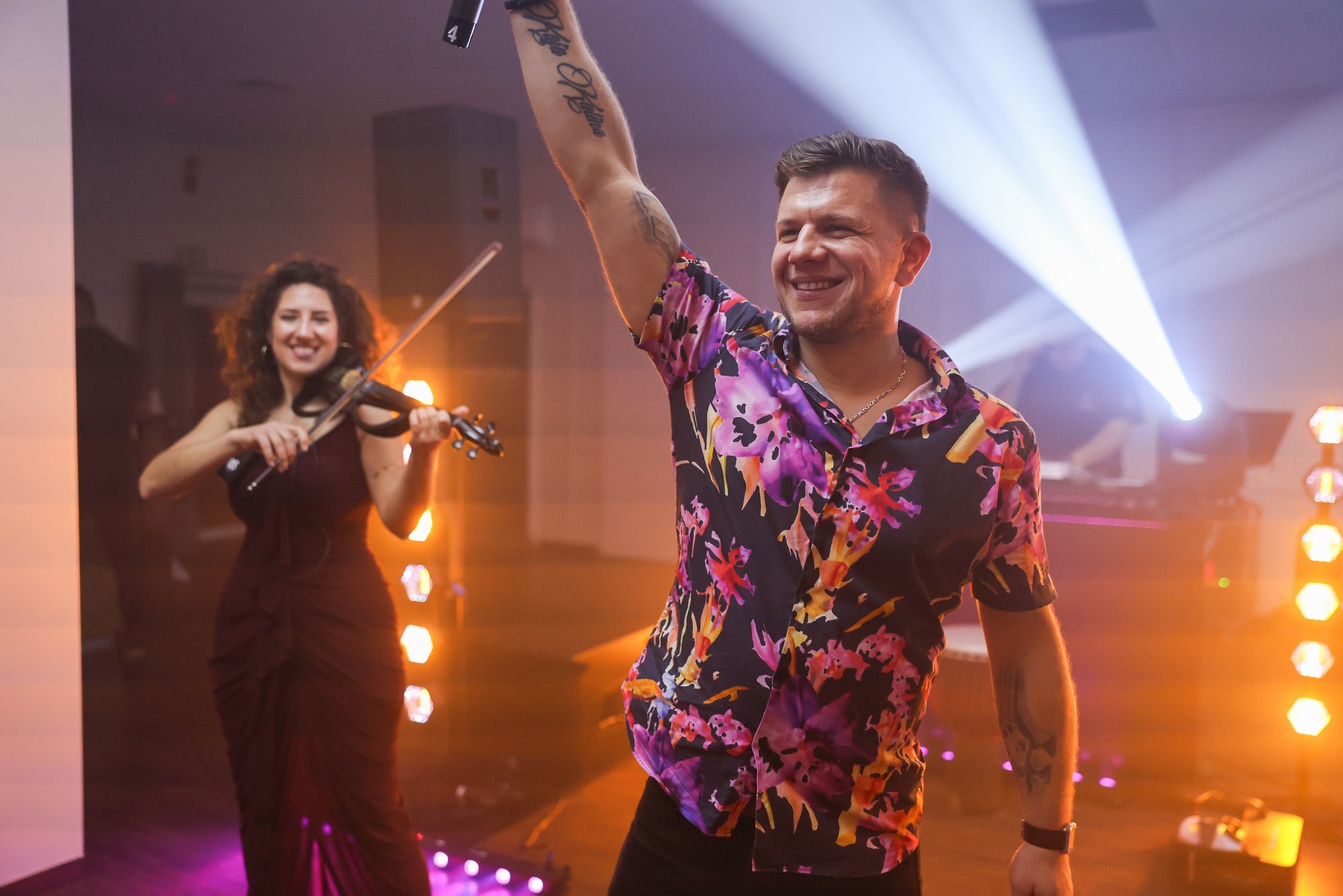 Uśmiechnięty mężczyzna w kwiecistej koszuli z uniesioną ręką, obok kobieta grająca na skrzypcach, oświetleni reflektorami na scenie.