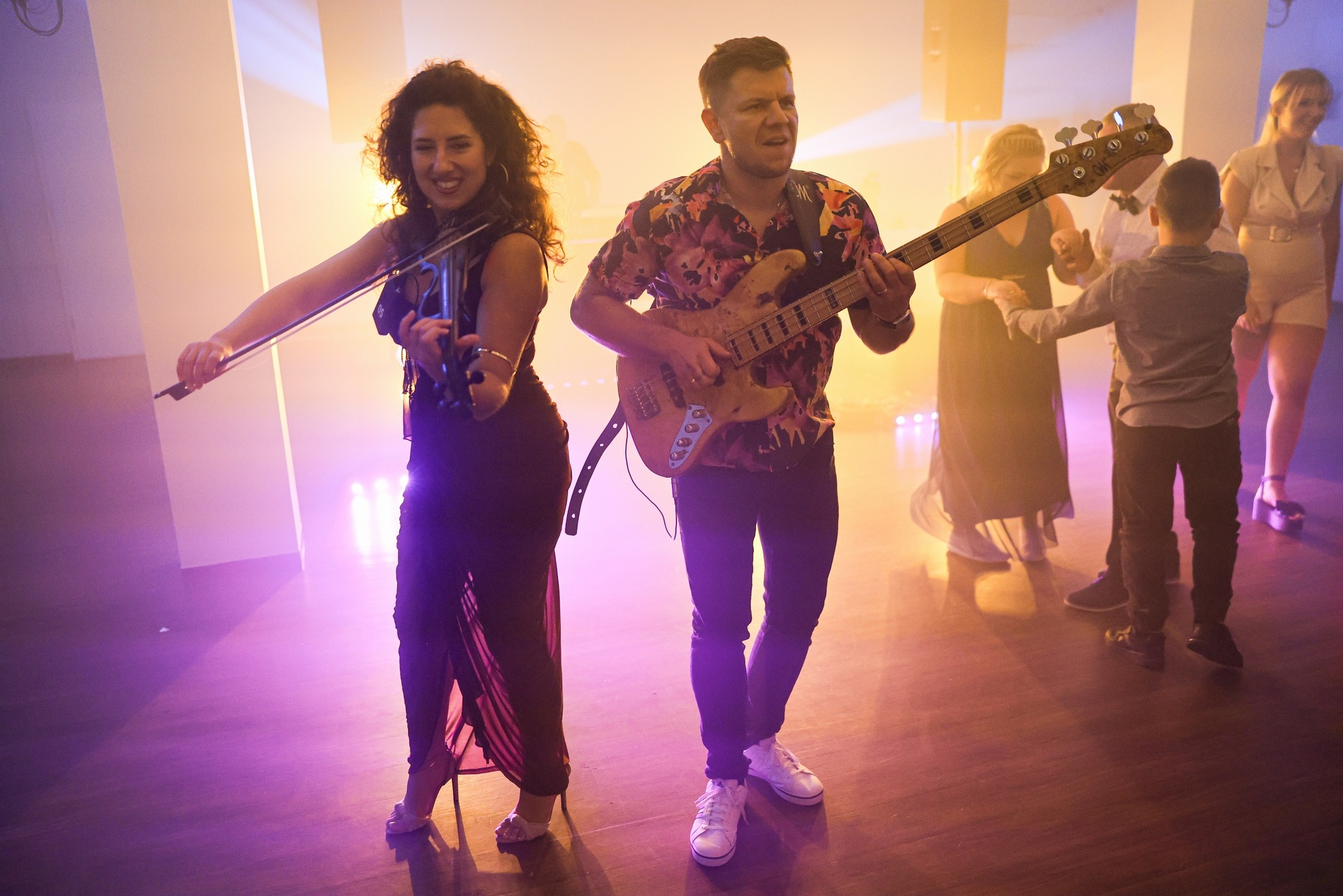 Duet muzyczny na scenie: kobieta gra na skrzypcach, mężczyzna na gitarze basowej. W tle tańczący goście w rozproszonym świetle.