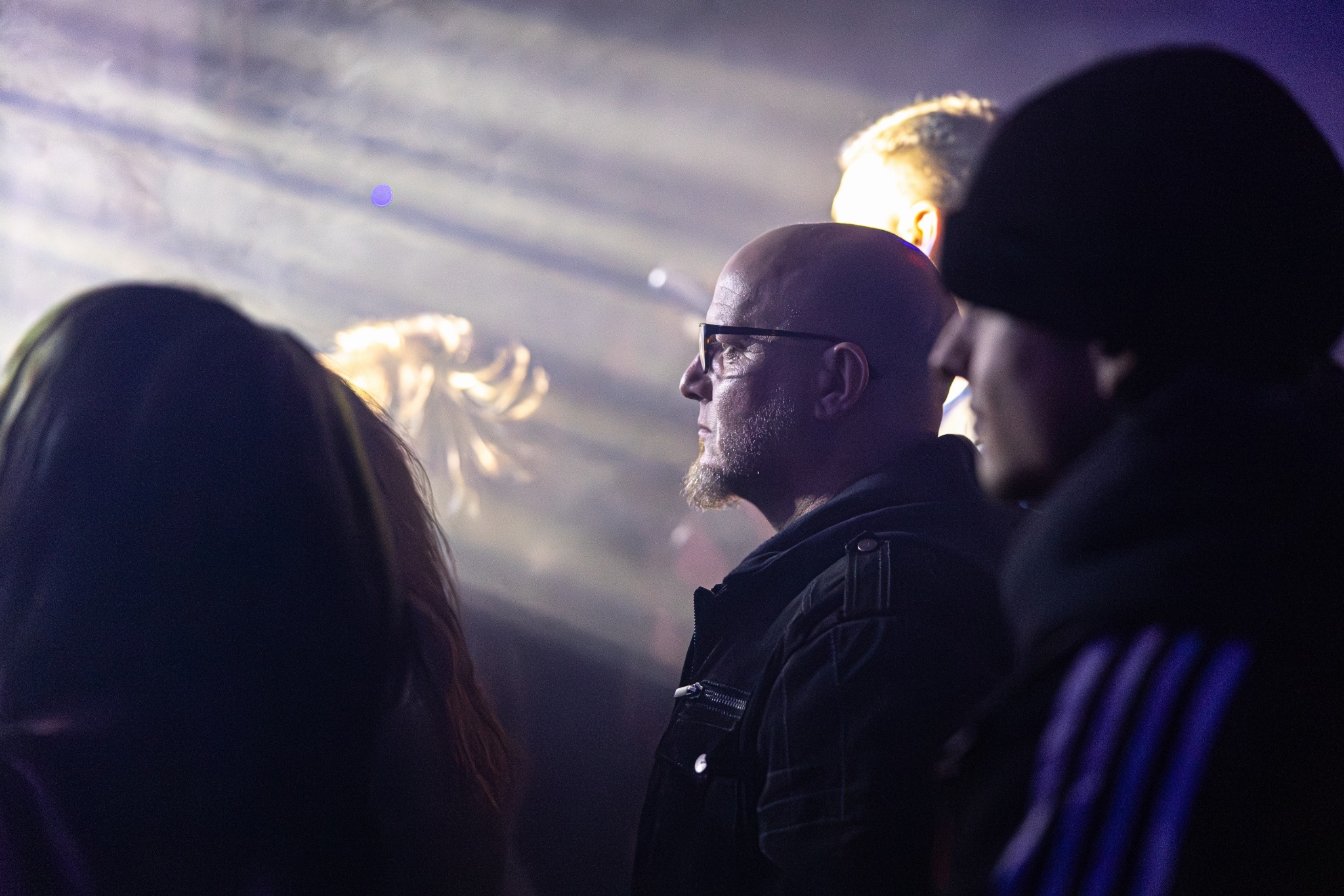 Mężczyzna w okularach z brodą, profil, w tłumie, oświetlony reflektorami, na imprezie. W tle rozmyte postacie i światła sceniczne. Ujęcie z dołu.