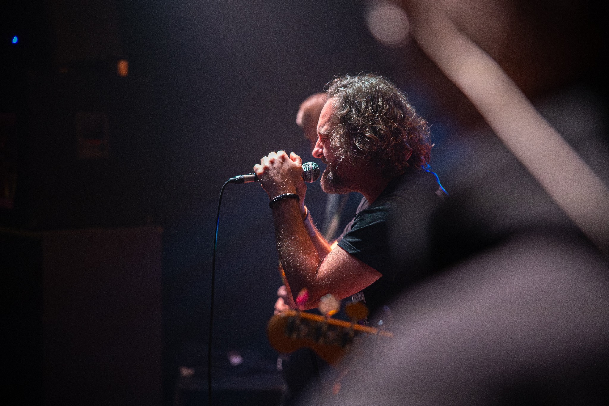 Mężczyzna z kręconymi włosami śpiewa do mikrofonu na scenie, w tle widoczny gitarzysta. Ujęcie z dołu, dynamiczne oświetlenie podkreśla ekspresję wokalisty.