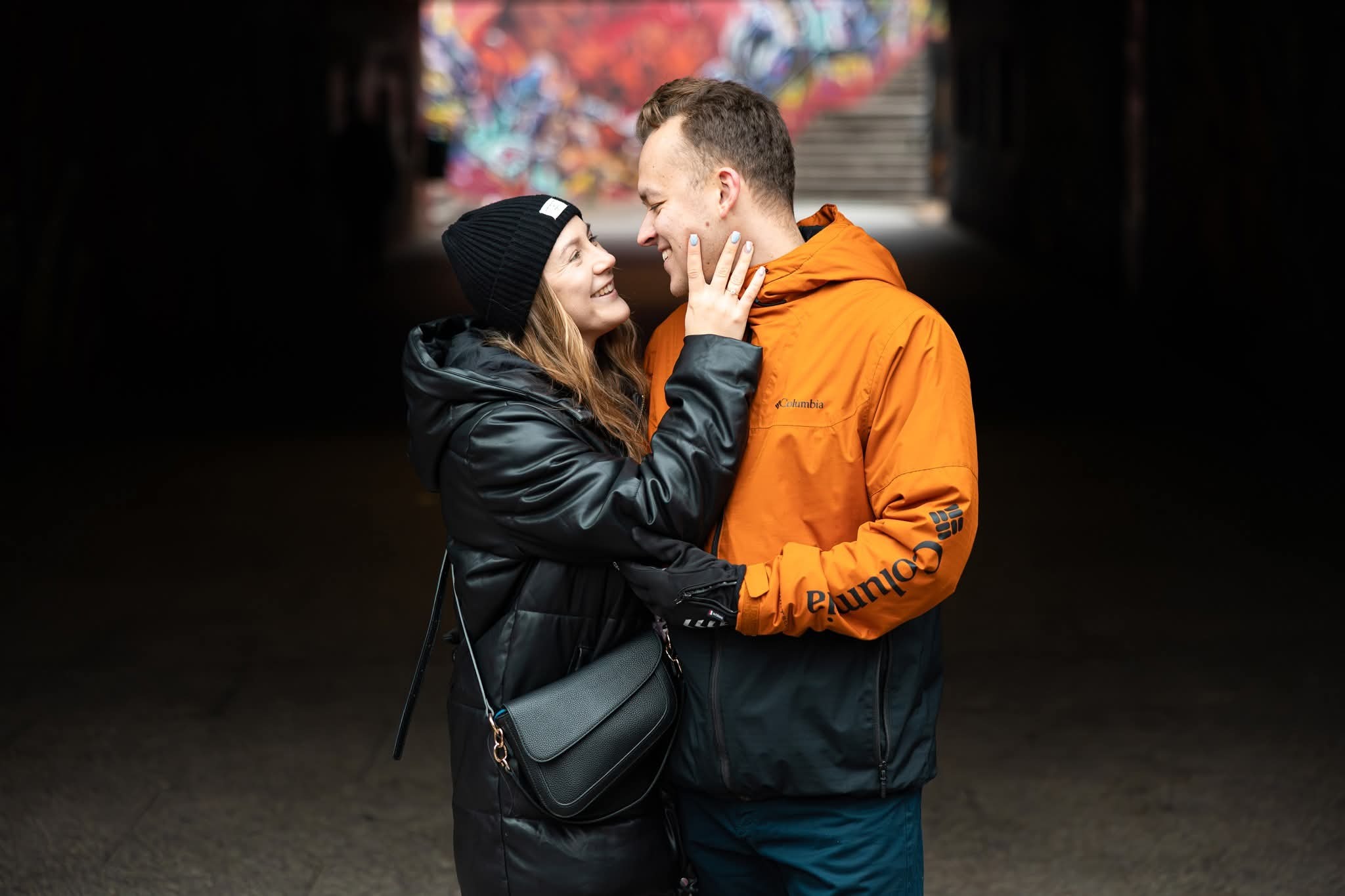 Zakochana para w tunelu z graffiti: kobieta w czapce i kurtce, mężczyzna w pomarańczowej kurtce Columbia, patrzą na siebie z uśmiechem. Romantyczna sesja narzeczeńska.