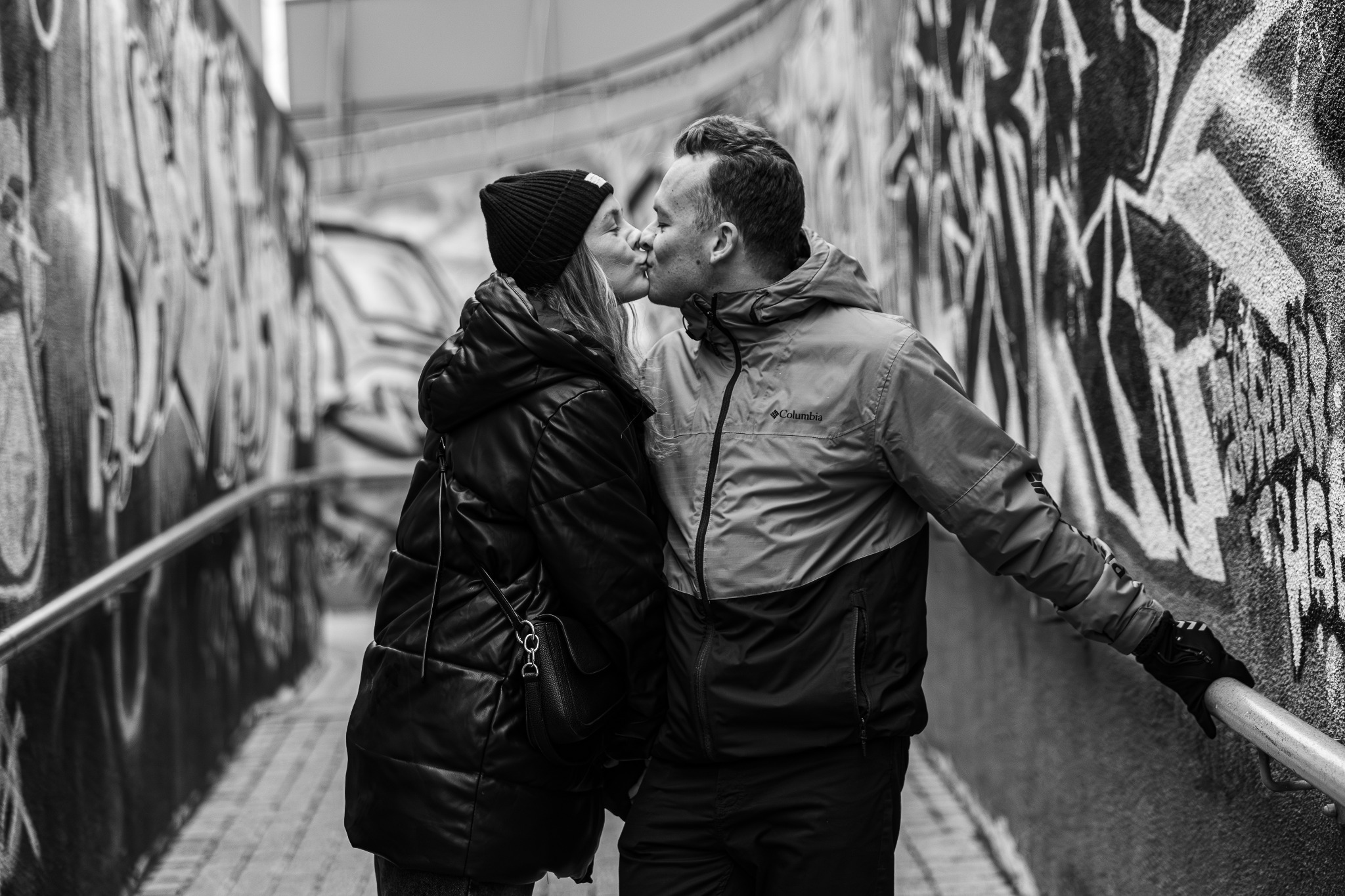 Czarno-białe zdjęcie całującej się pary w tunelu z graffiti. Kobieta w czapce i kurtce, mężczyzna w kurtce Columbia. Ujęcie z boku, blisko, z widocznymi detalami.
