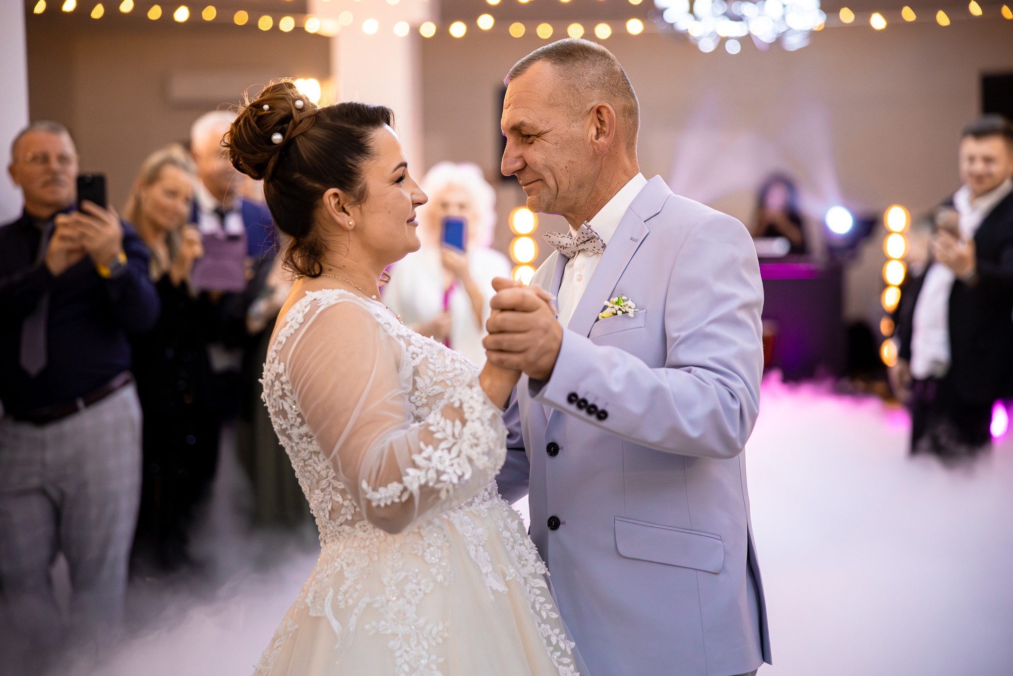 Para w strojach ślubnych tańczy w obłokach dymu, oświetlona ciepłymi światłami. Goście obserwują i robią zdjęcia.