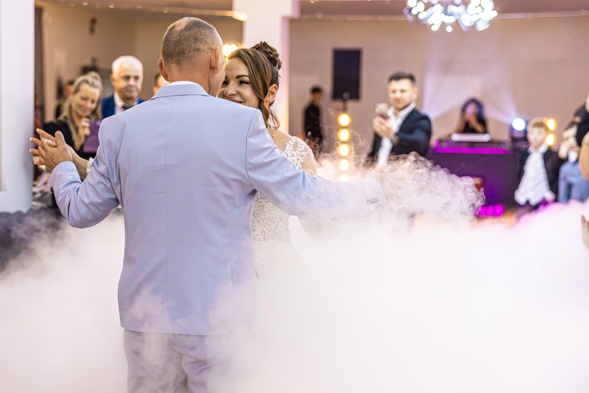 Para młoda w tańcu, otoczona efektem dymu na parkiecie. Pan młody w błękitnym garniturze, panna młoda w białej sukni. Goście obserwują w tle.