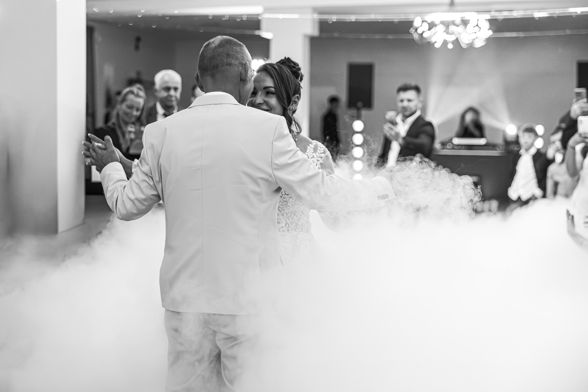 Czarno-białe zdjęcie pary młodej w tańcu, otoczonej mgłą, z gośćmi w tle. Panna młoda uśmiecha się, patrząc na partnera. Scena z wesela.