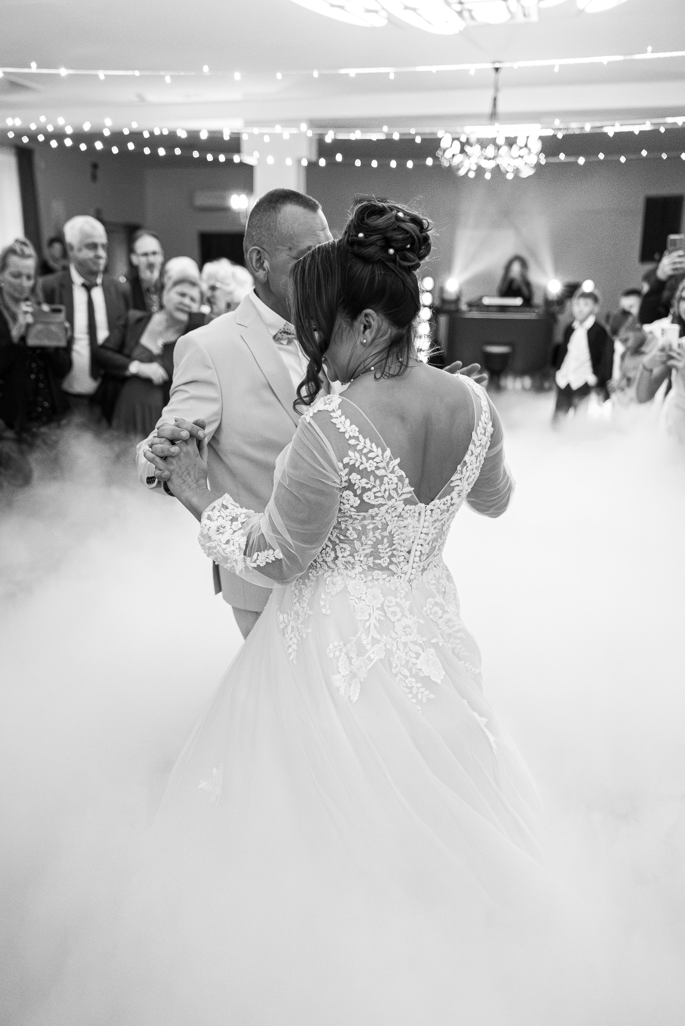 Czarno-białe zdjęcie pary młodej podczas pierwszego tańca w dymie, z gośćmi w tle. Suknia panny młodej z koronkowymi detalami na plecach, garnitur pana młodego w jasnym odcieniu.