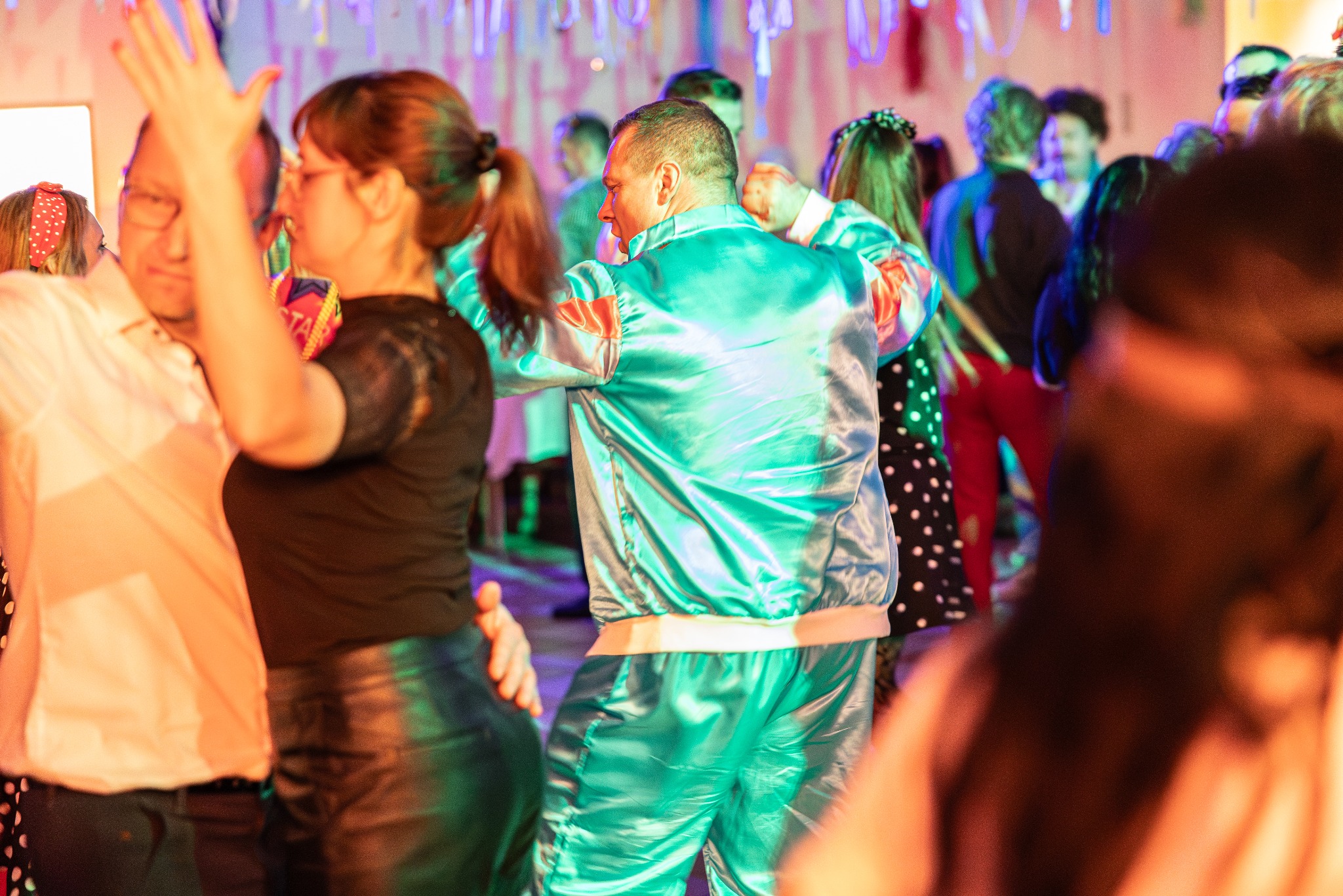 Roztańczony tłum w karnawałowych strojach, mężczyzna w błyszczącym turkusowym dresie w centrum kadru, imprezowa atmosfera, kolorowe światła i dekoracje.