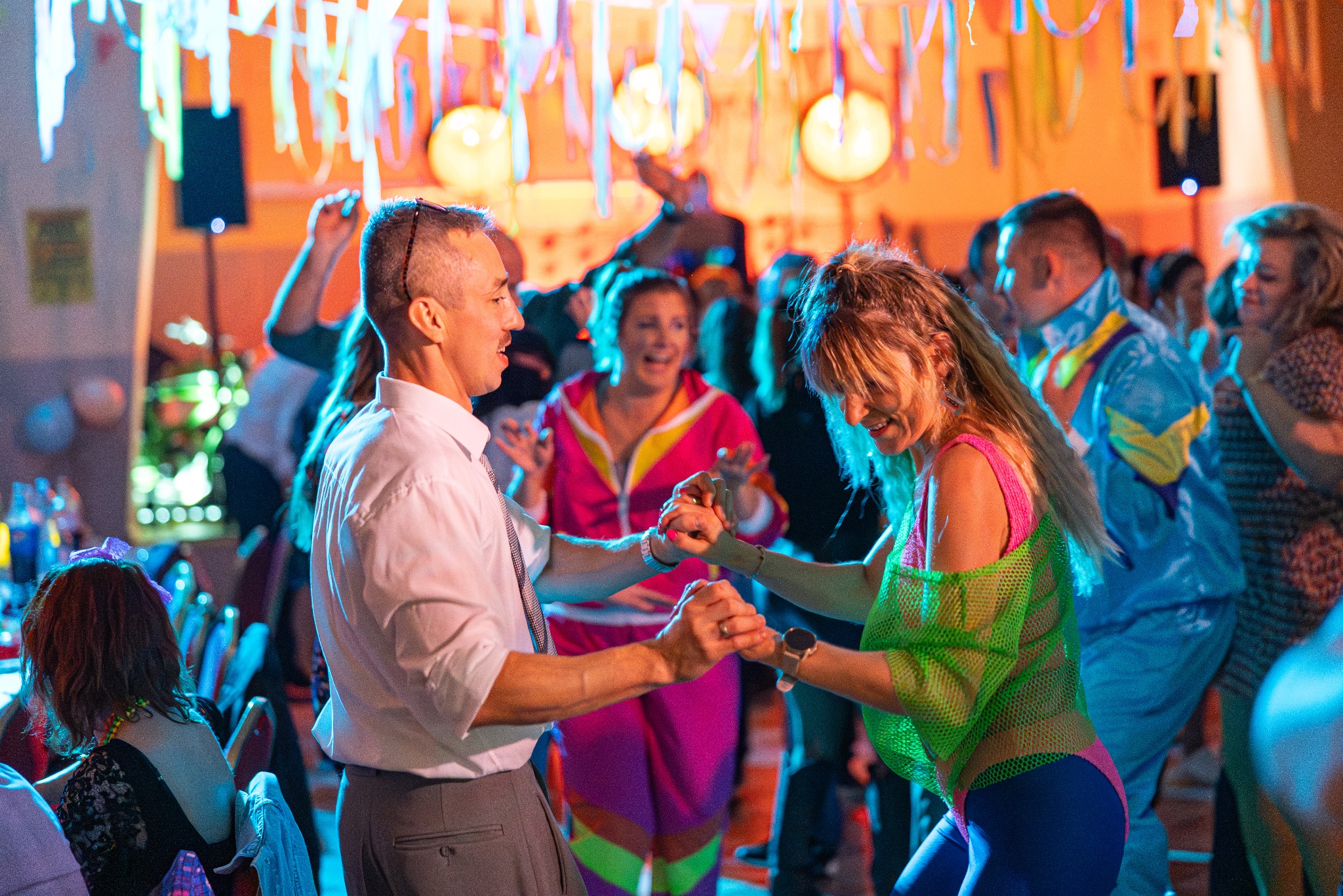 Para w strojach retro tańczy na parkiecie podczas imprezy tematycznej. Kolorowe dekoracje i rozbawieni goście w tle tworzą radosną atmosferę.