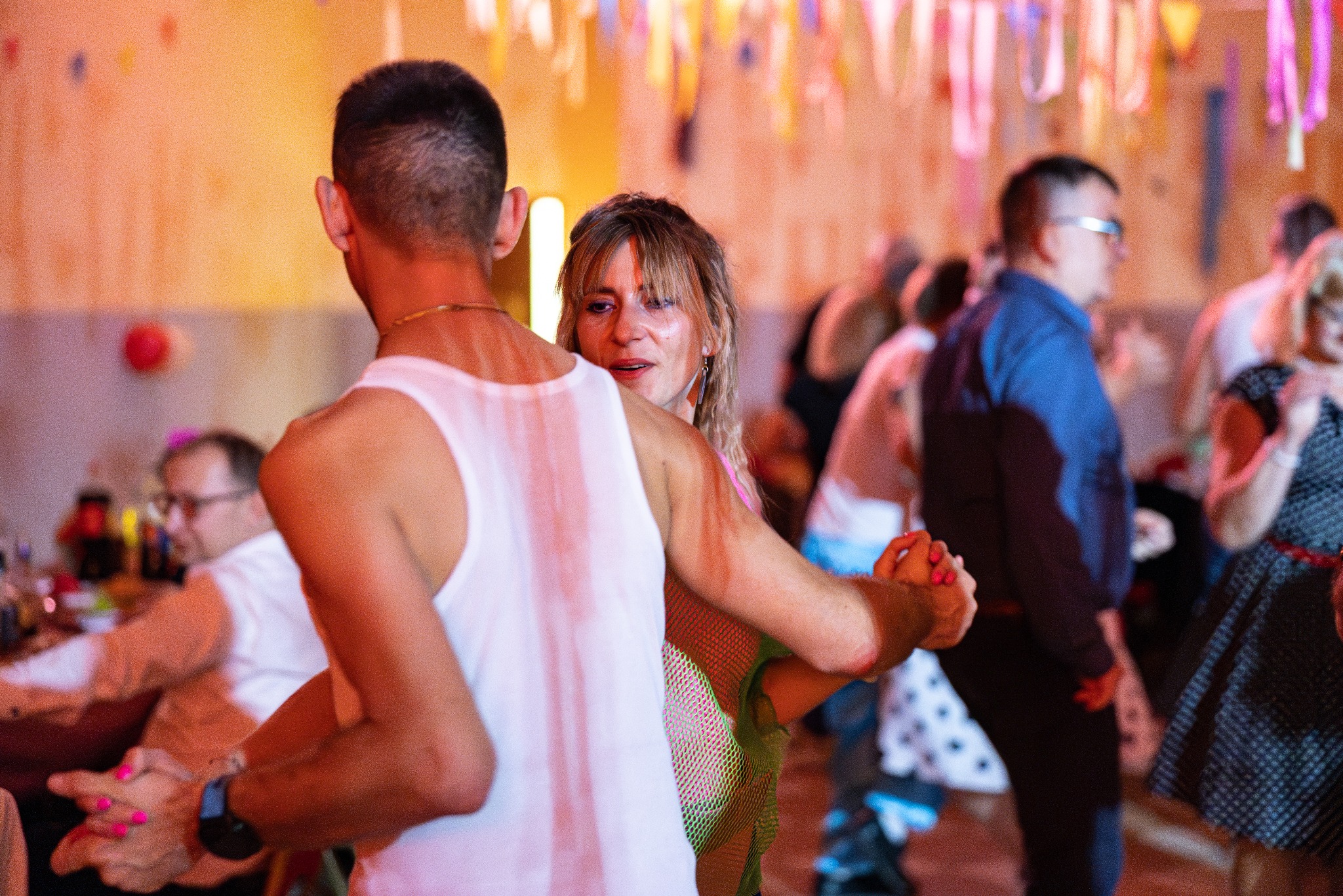 Para tańczy na parkiecie podczas imprezy. Kobieta patrzy w obiektyw, mężczyzna w białym podkoszulku odwrócony tyłem. W tle dekoracje i inni goście.