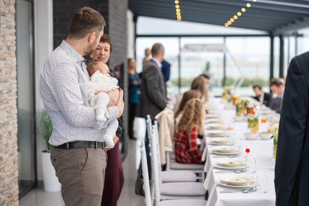 Mężczyzna trzyma na rękach niemowlę w białej sukience na tle sali bankietowej z gośćmi i długim stołem. Ujęcie z boku, z widocznymi detalami stroju dziecka.