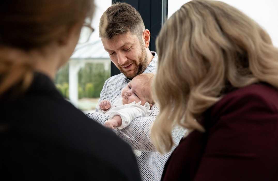 Tata trzymający niemowlę na rękach, z uśmiechem patrzy na dziecko. W tle widoczne dwie osoby, prawdopodobnie rodzina, obserwujące tę wzruszającą chwilę.