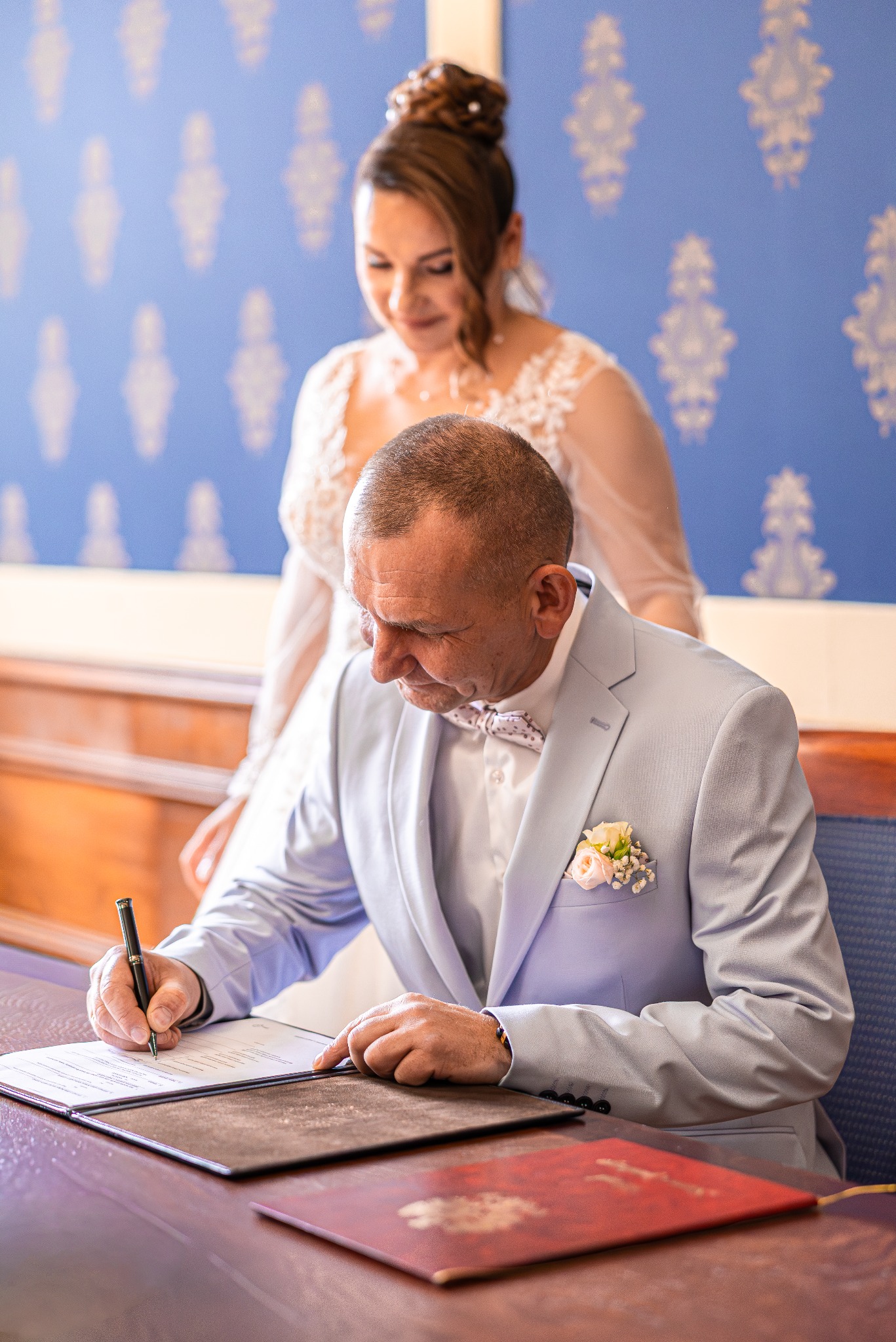 Pan młody w błękitnym garniturze podpisuje akt ślubu, panna młoda w białej sukni obserwuje. Uroczysta chwila w eleganckim wnętrzu.