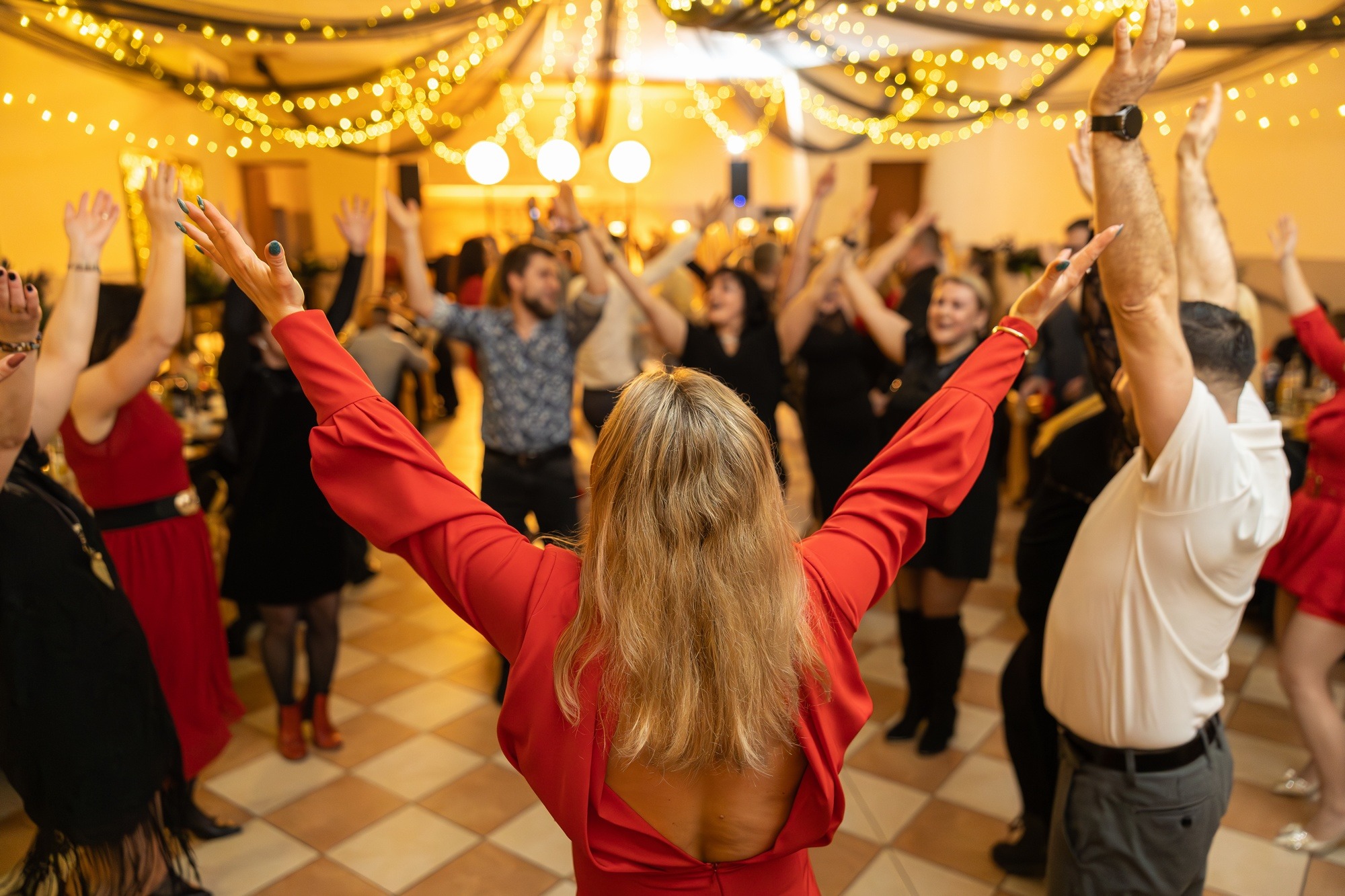 Roztańczona grupa ludzi z podniesionymi rękami, widok zza kobiety w czerwonej sukience na parkiecie w ciepłym oświetleniu podczas imprezy.