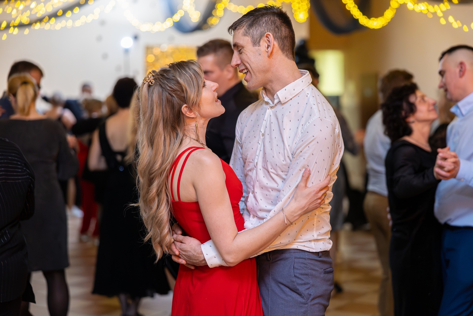 Para w objęciach na parkiecie, kobieta w czerwonej sukience, mężczyzna w białej koszuli. W tle tańczący goście i dekoracje świetlne.