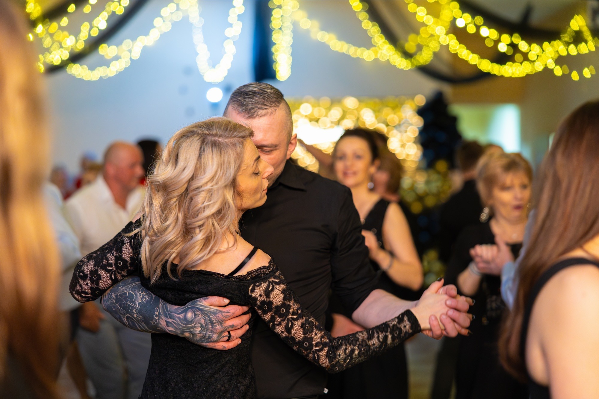 Para w czarnych strojach tańczy na parkiecie podczas eventu, oświetlonym girlandami. Mężczyzna ma tatuaż na ramieniu. W tle widoczni inni goście.