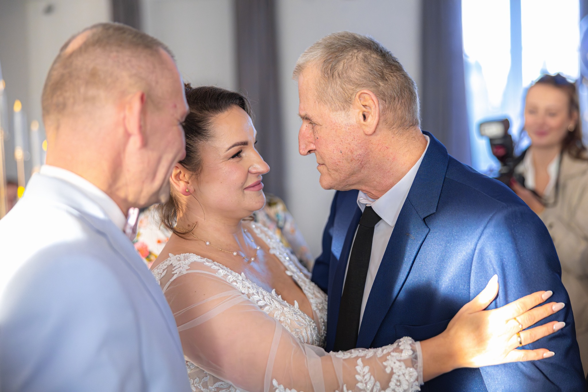 Uśmiechnięta panna młoda w koronkowej sukni spogląda na pana młodego w granatowym garniturze. W tle fotografka z aparatem.