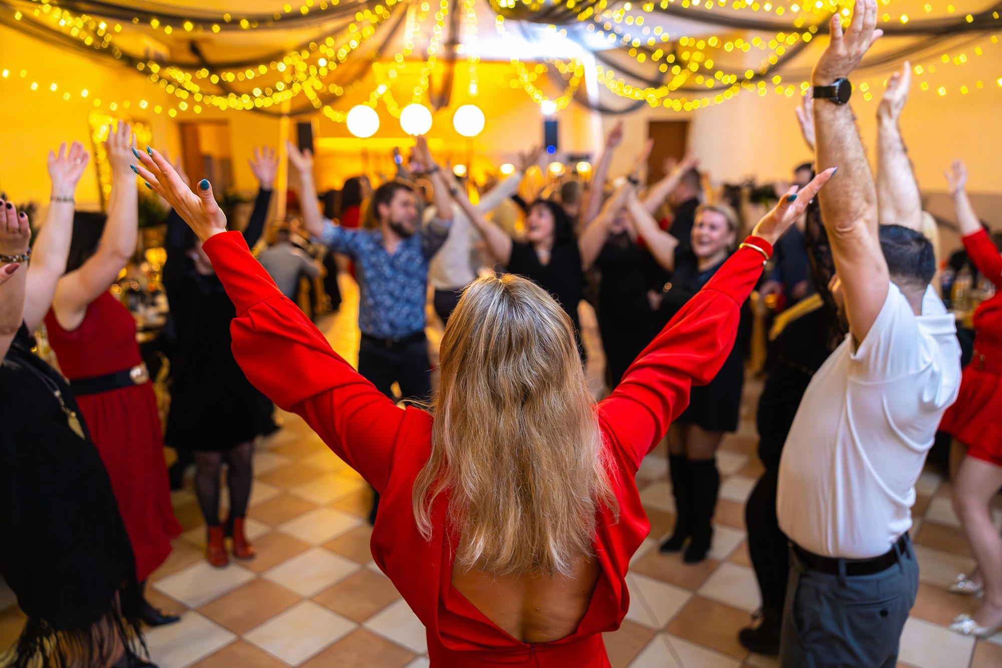 Roztańczony tłum z uniesionymi rękami, widok zza kobiety w czerwonej sukience, ciepłe światło girland, parkiet w kratkę, radosna atmosfera.