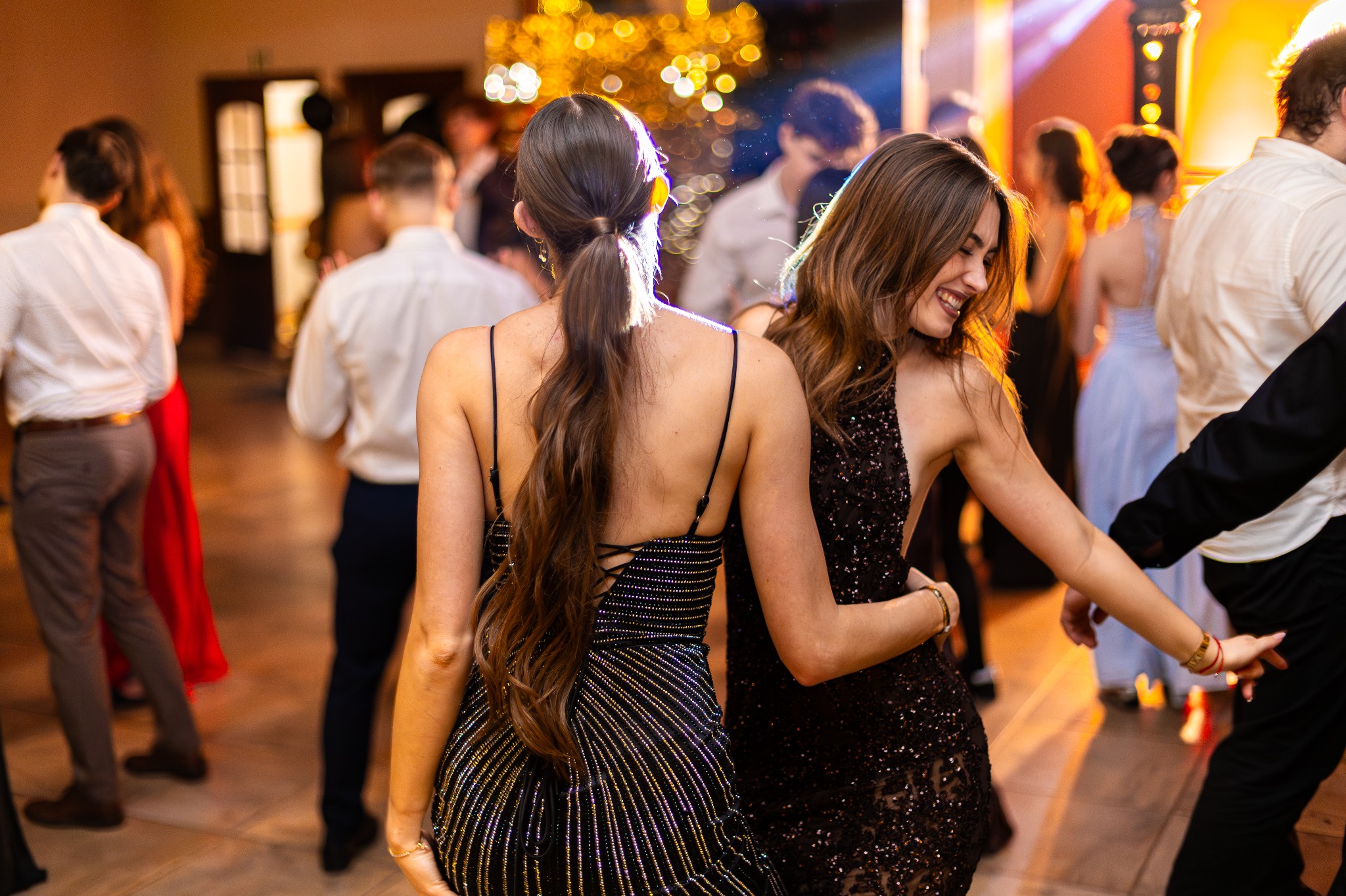 Dwie uśmiechnięte kobiety w błyszczących sukienkach na parkiecie podczas eventu. W tle rozmyte postacie i dekoracje świetlne.