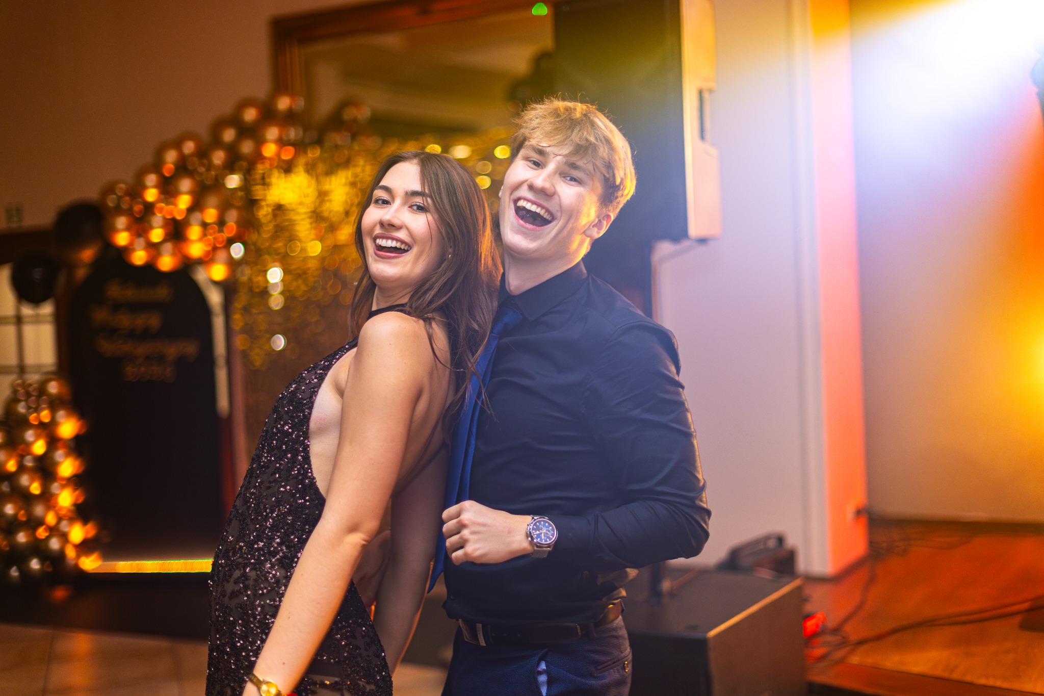 Uśmiechnięta para tańczy na parkiecie podczas eventu. Kobieta w cekinowej sukience, mężczyzna w koszuli i krawacie. W tle dekoracje świetlne i nagłośnienie.