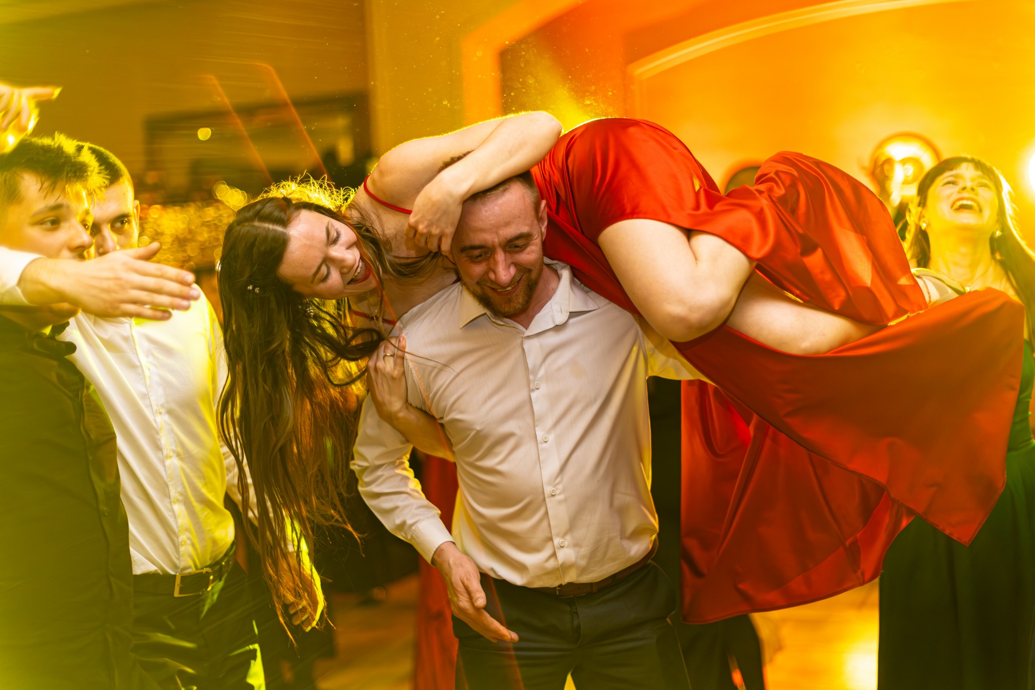 Radosna zabawa weselna w Głogowie: mężczyzna trzyma na ramionach uśmiechniętą kobietę w czerwonej sukni, otoczonych przez rozbawionych gości w ciepłym oświetleniu.