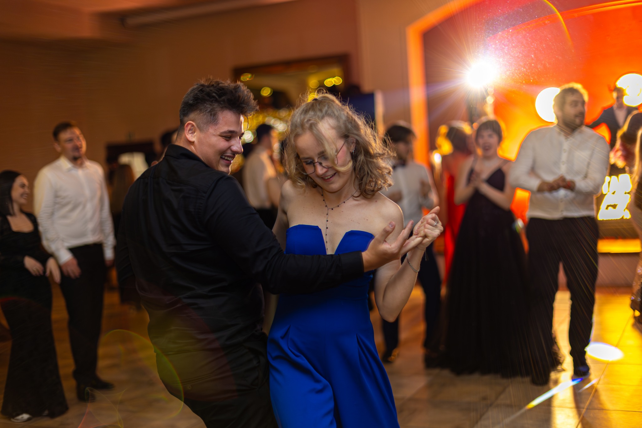Para tańczy na parkiecie podczas eventu. Kobieta w niebieskiej sukience, mężczyzna w czarnej koszuli. W tle rozświetlona scena i goście.
