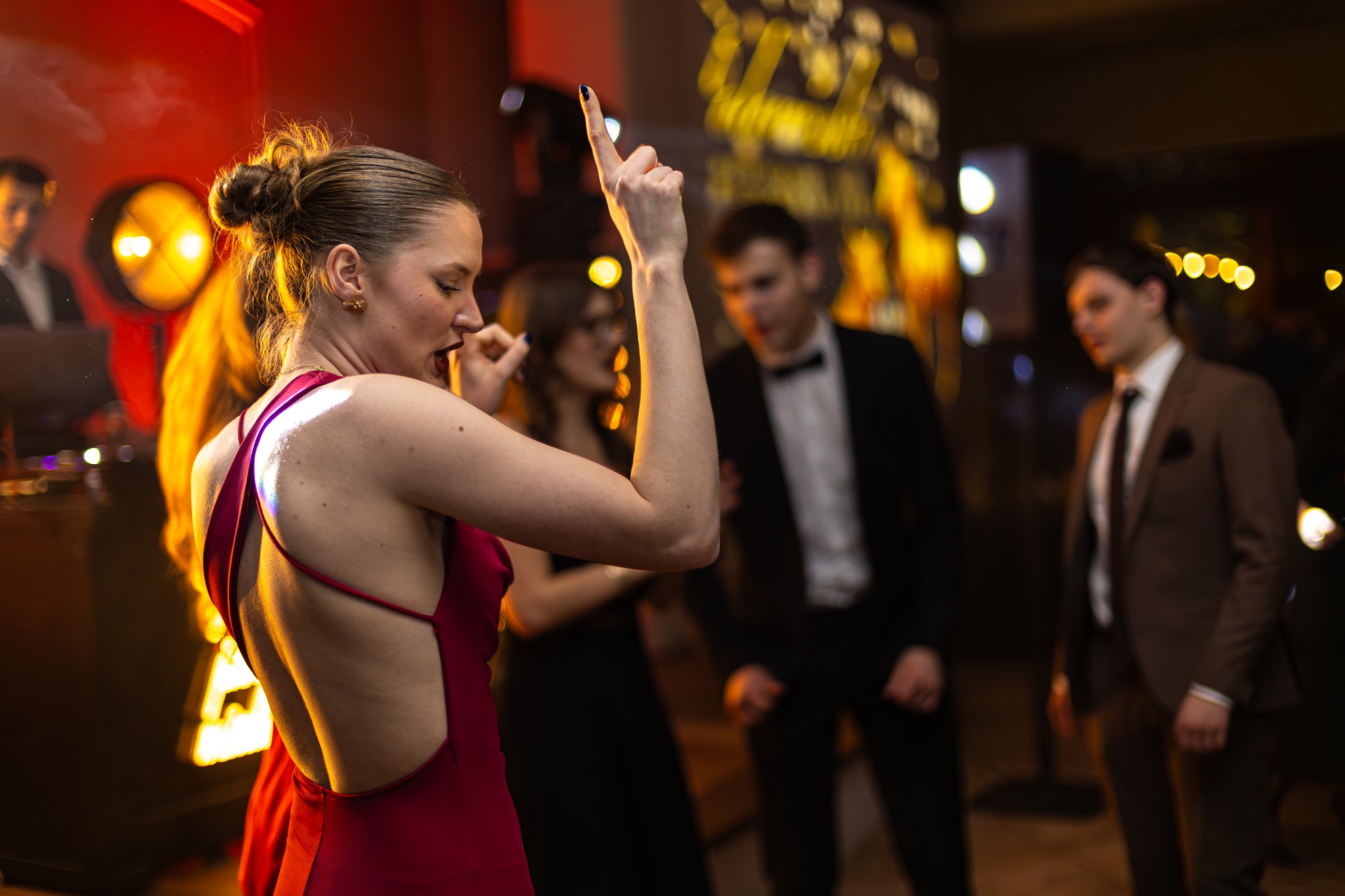 Kobieta w czerwonej sukni tańczy na parkiecie podczas eventu. W tle inni goście w strojach wieczorowych i dekoracje świetlne.