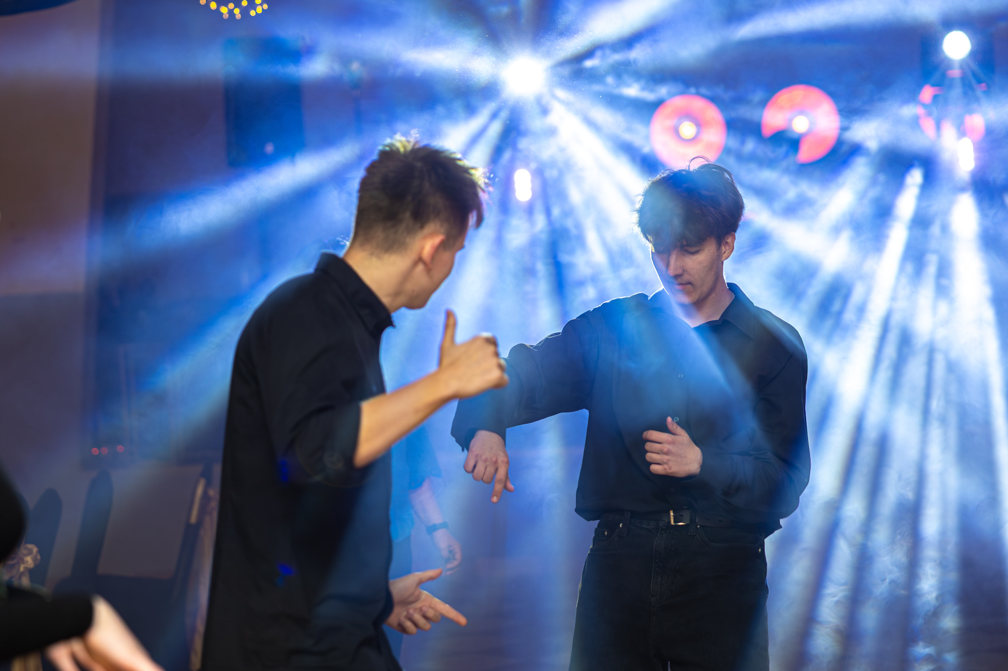 Dwóch młodych mężczyzn w czarnych koszulach pozuje na tle niebieskich świateł podczas eventu. Jeden pokazuje kciuk w górę, drugi patrzy w dół. Dynamiczne oświetlenie.