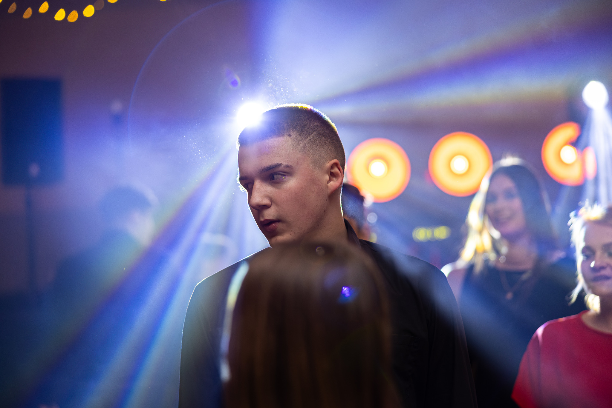 Młody mężczyzna z modną fryzurą na imprezie, oświetlony reflektorami. W tle rozmazane sylwetki gości i dekoracyjne światła. Dynamiczne ujęcie z bliska.