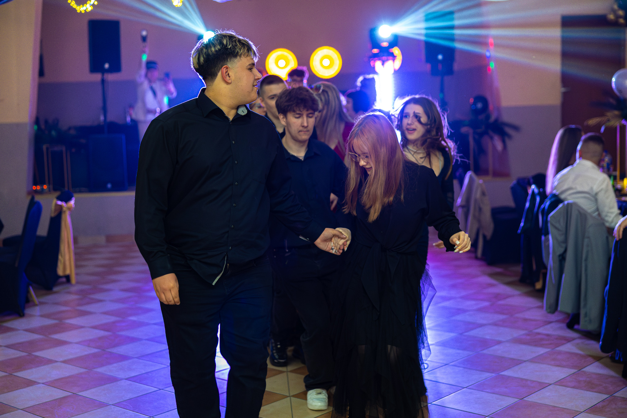 Grupa młodych ludzi w ciemnych strojach, trzymających się za ręce, idzie w kierunku światła na parkiecie podczas imprezy. W tle widoczna scena i oświetlenie.