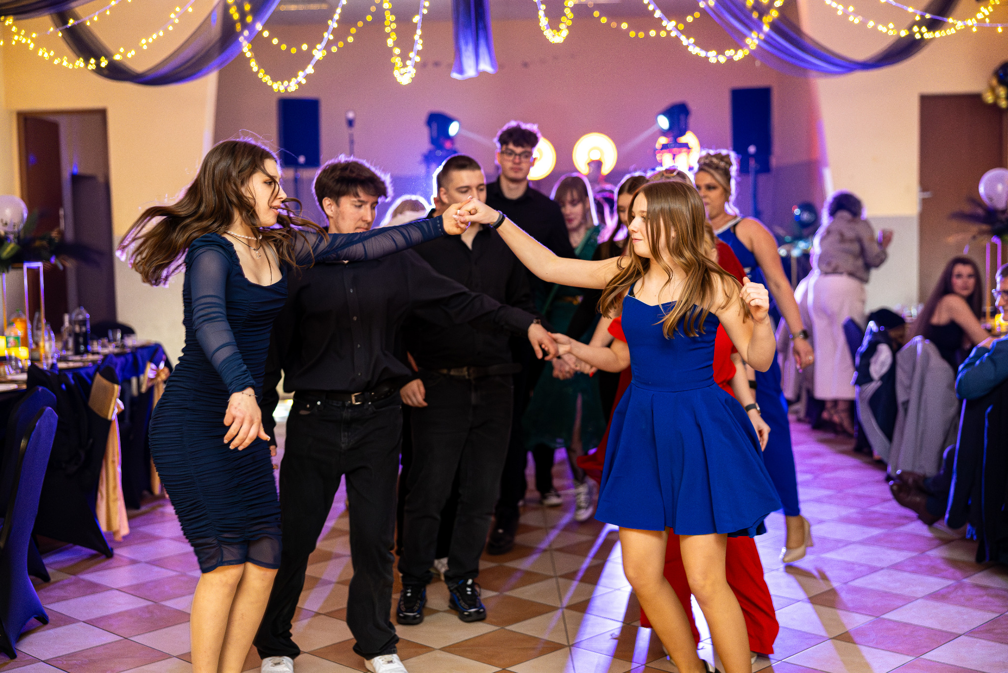 Roztańczona grupa młodych ludzi na parkiecie podczas eventu w Głogowie. Dekoracje świetlne i eleganckie stroje tworzą radosną atmosferę.