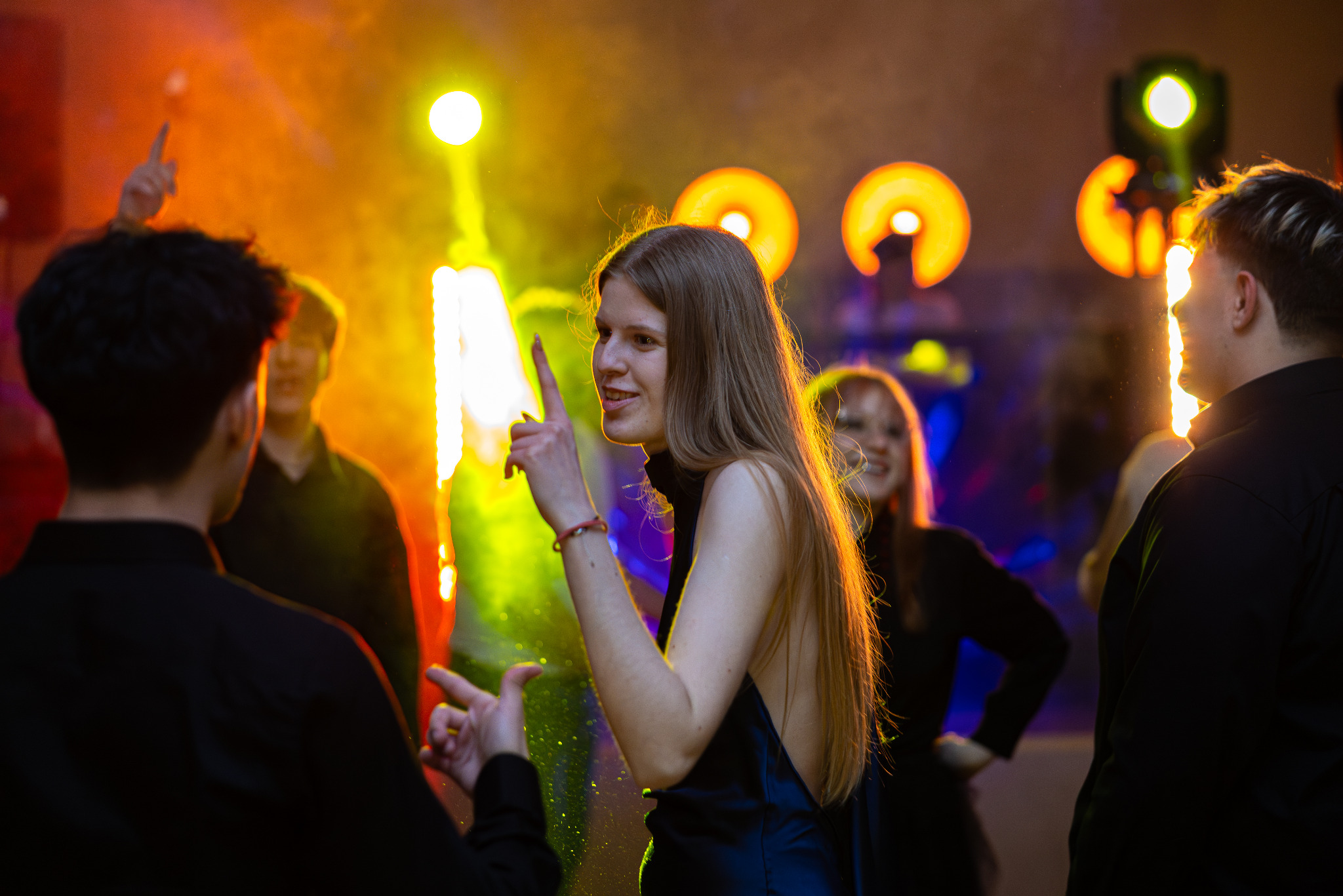 Rozświetlona scena z młodą kobietą w centrum, unoszącą palec. W tle rozmazane sylwetki i kolorowe światła tworzą dynamiczną atmosferę imprezy.