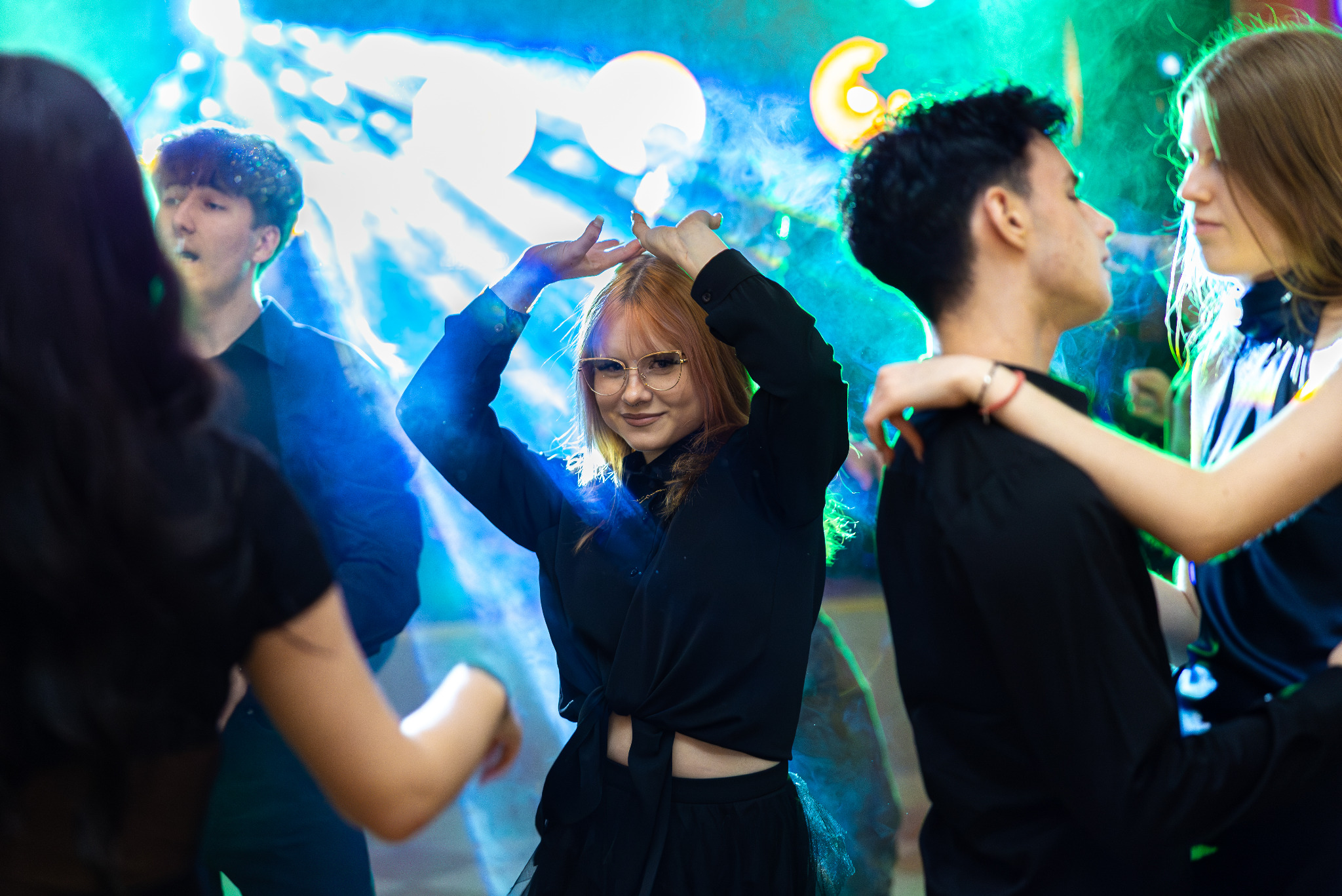 Młodzi ludzie tańczą w klubie, oświetleni kolorowymi światłami. Dziewczyna w okularach uśmiecha się, unosząc ręce. Para w objęciach, w tle inne osoby.