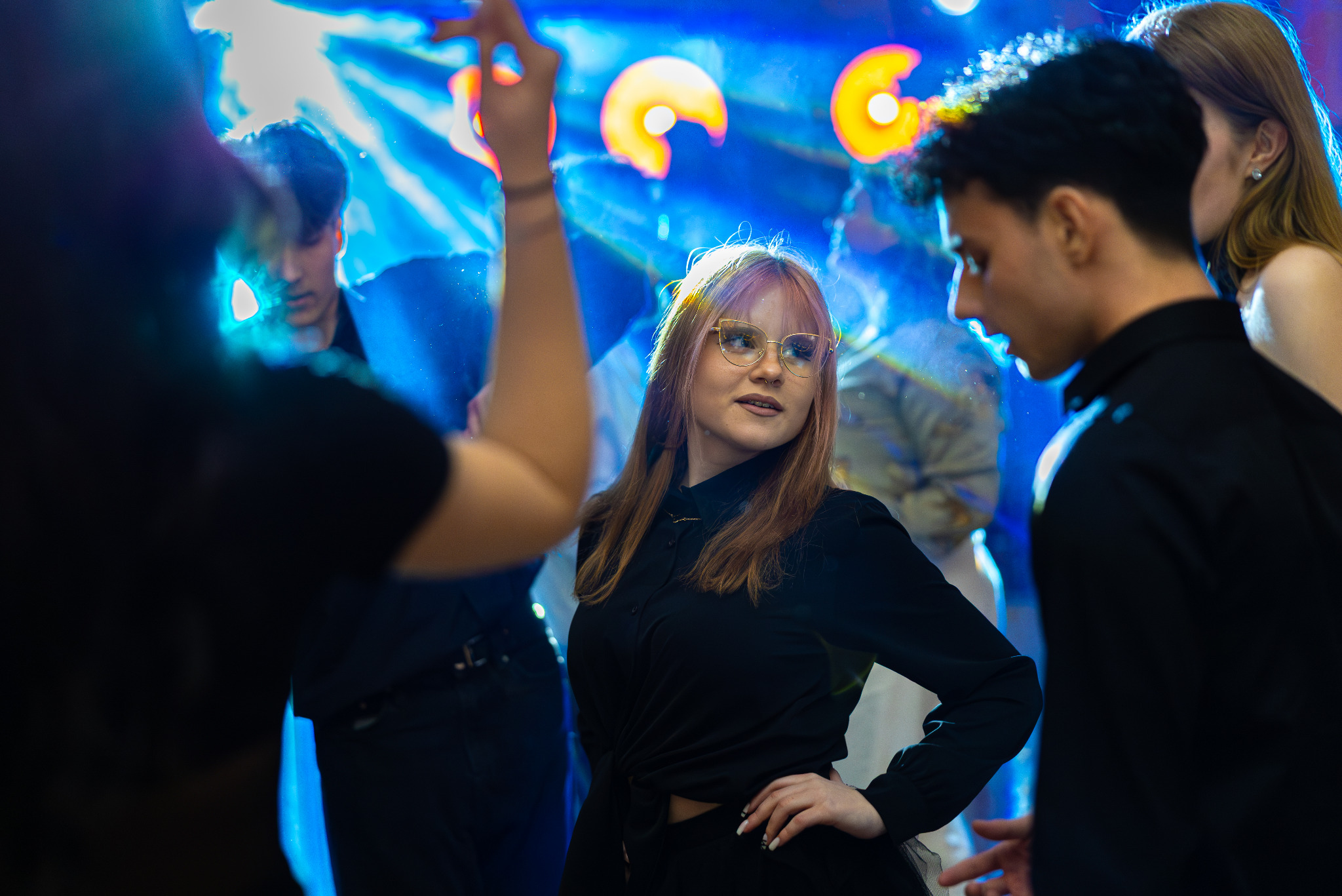 Młoda kobieta w okularach tańczy na parkiecie w otoczeniu innych osób, oświetlona niebieskim światłem i reflektorami. Ujęcie z dołu, dynamiczna scena.