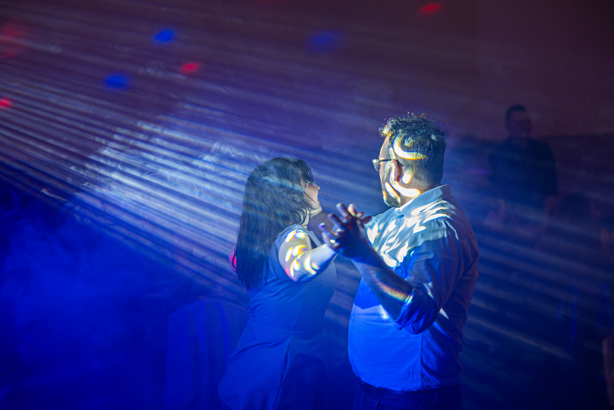 Para tańczy w niebieskim świetle reflektorów na evencie. Mężczyzna w okularach i białej koszuli, kobieta w sukience. W tle zamazana postać.