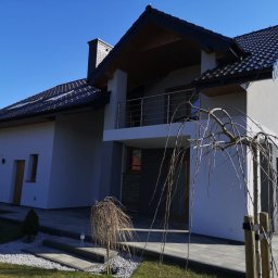 Kamil Cichecki Usługi Remontowo - Wykończeniowe - Nowoczesny dom jednorodzinny z jasnoszarą i ciemnoszarą elewacją, ciemnym dachem z dachówek, balkonem z metalową balustradą. Wokół kostka brukowa i bezlistne drzewko ozdobne.