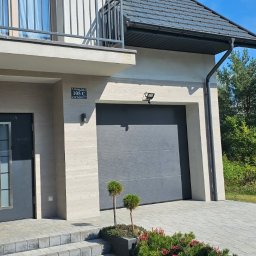Kamil Cichecki Usługi Remontowo - Wykończeniowe - Nowoczesna fasada domu z numerem 105 C, szarą bramą garażową i drzwiami wejściowymi z pionowymi przeszkleniami. Jasna elewacja z gładkich tynków i drewnopodobnych paneli, brukowany podjazd i balkon...