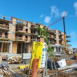 PFB PIOTR FIEDORUK - Wielopiętrowy budynek w trakcie budowy z cegły i betonu, widoczny dźwig, rusztowania, tablica informacyjna i pracownicy w odblaskowych kamizelkach. Błękitne niebo w tle.