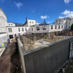 PFB PIOTR FIEDORUK - Budowa budynku żelbetowego w Kołobrzegu: widok na zbrojenie ścian fundamentowych z perspektywy placu budowy, z pracownikami w tle i dźwigiem w górnej części kadru.