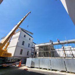 PFB PIOTR FIEDORUK - Montaż stropu budynku z użyciem dźwigu, widoczne rusztowania i pracownicy. Ujęcie z dołu, błękitne niebo w tle. Dźwig firmy Dźwigi-Kołobrzeg.