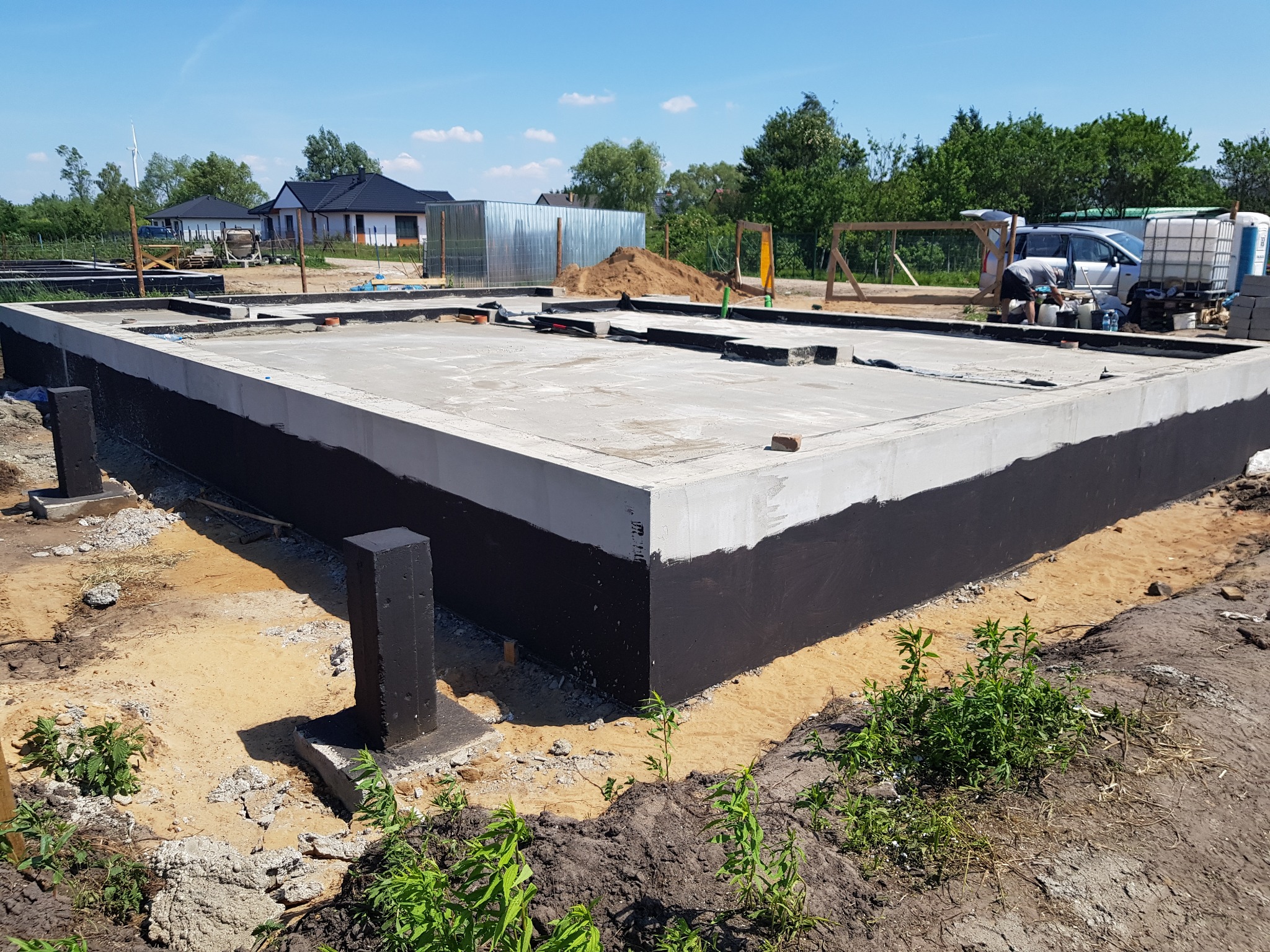 Zalana płyta fundamentowa z izolacją pionową, widoczne czarne słupy fundamentowe. W tle zabudowa jednorodzinna i teren budowy w słoneczny dzień.