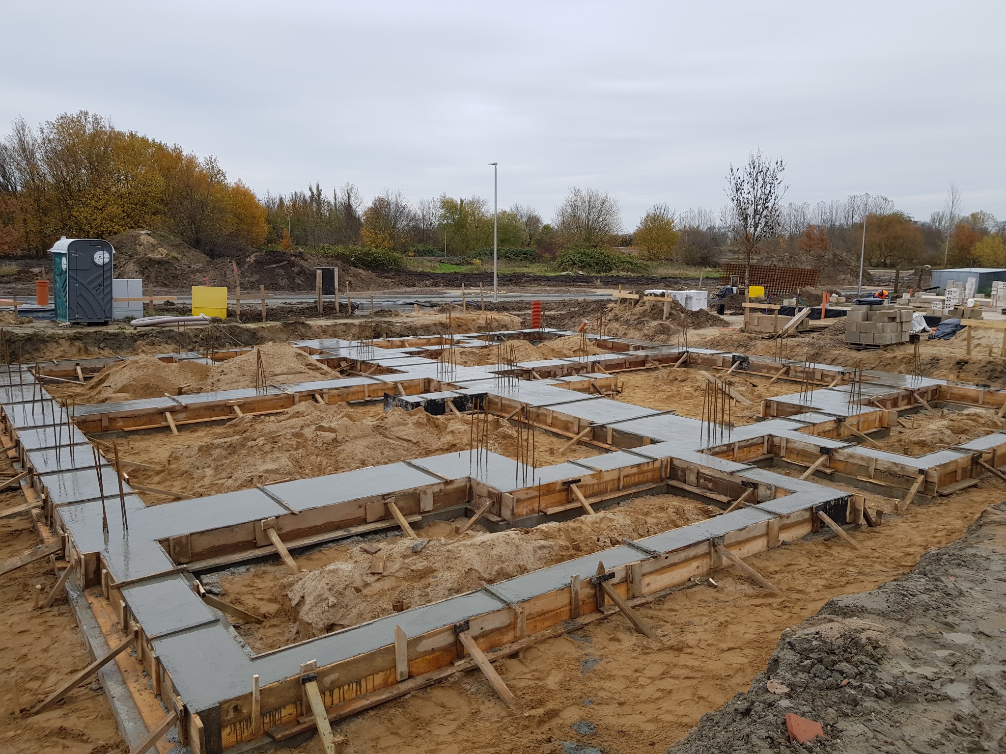 Zalane betonem fundamenty domu jednorodzinnego w drewnianym szalunku, wystające pręty zbrojeniowe, teren budowy z widoczną toaletą przenośną i drzewami w tle.