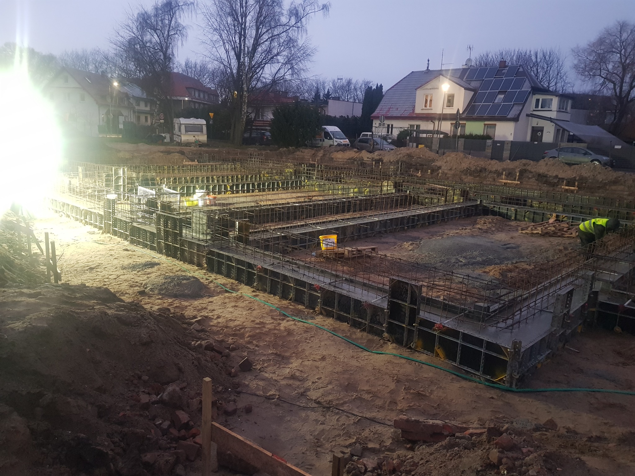 Zbrojenie fundamentów domu jednorodzinnego w trakcie budowy, widoczne szalunki i stalowe pręty, oświetlone reflektorem, w tle zabudowa mieszkalna.