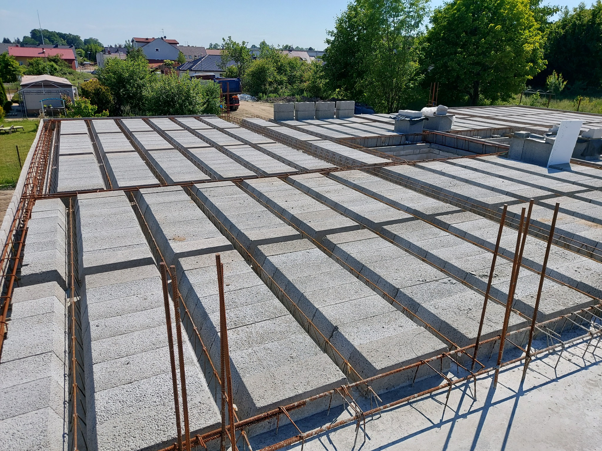 Zbrojona konstrukcja stropu z betonowych pustaków szalunkowych, widok z góry, w tle zabudowa jednorodzinna i zieleń. Etap budowy domu.