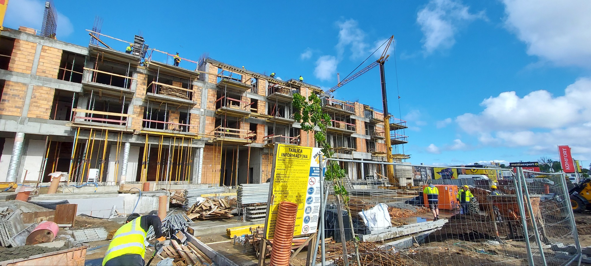 Wielopiętrowy budynek w trakcie budowy z cegły i betonu, widoczny dźwig, rusztowania, tablica informacyjna i pracownicy w odblaskowych kamizelkach. Błękitne niebo w tle.
