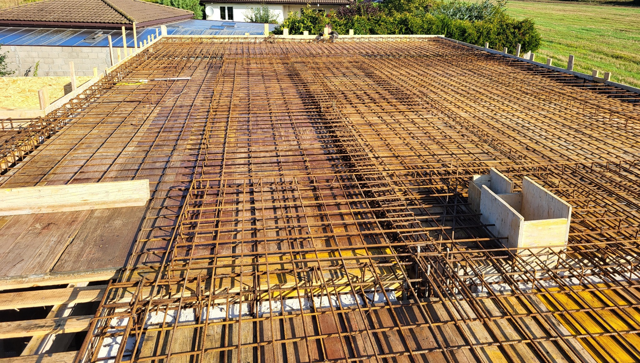 Siatka zbrojeniowa na deskowaniu, przygotowana do wylewki betonowej. Widoczne elementy szalunków i konstrukcji nośnej. W tle zieleń i zabudowa jednorodzinna.