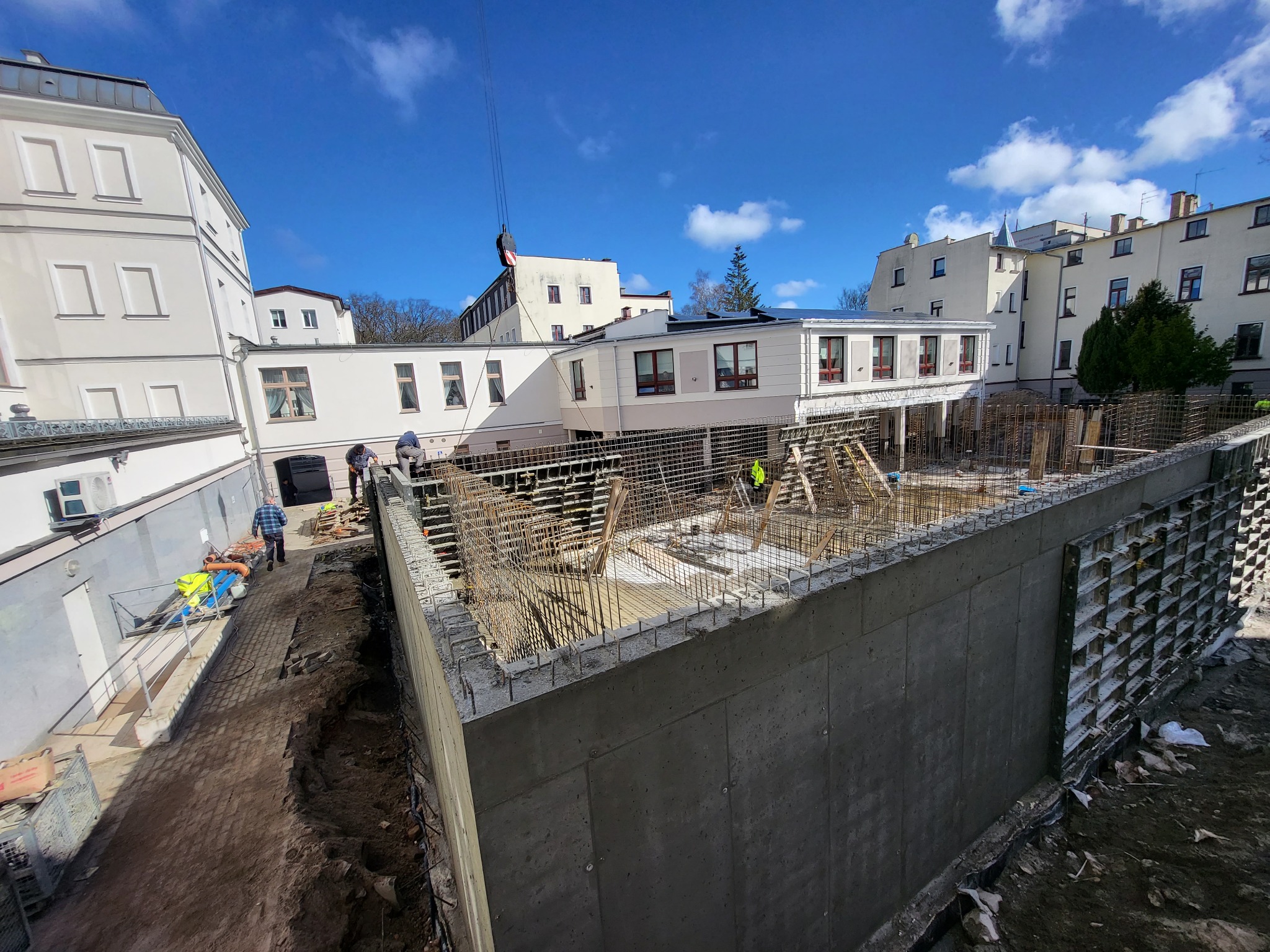 Budowa budynku żelbetowego w Kołobrzegu: widok na zbrojenie ścian fundamentowych z perspektywy placu budowy, z pracownikami w tle i dźwigiem w górnej części kadru.